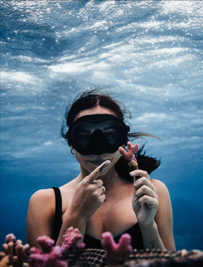 A freediver just under the surface, holding a pink coral fragment up beside her mask