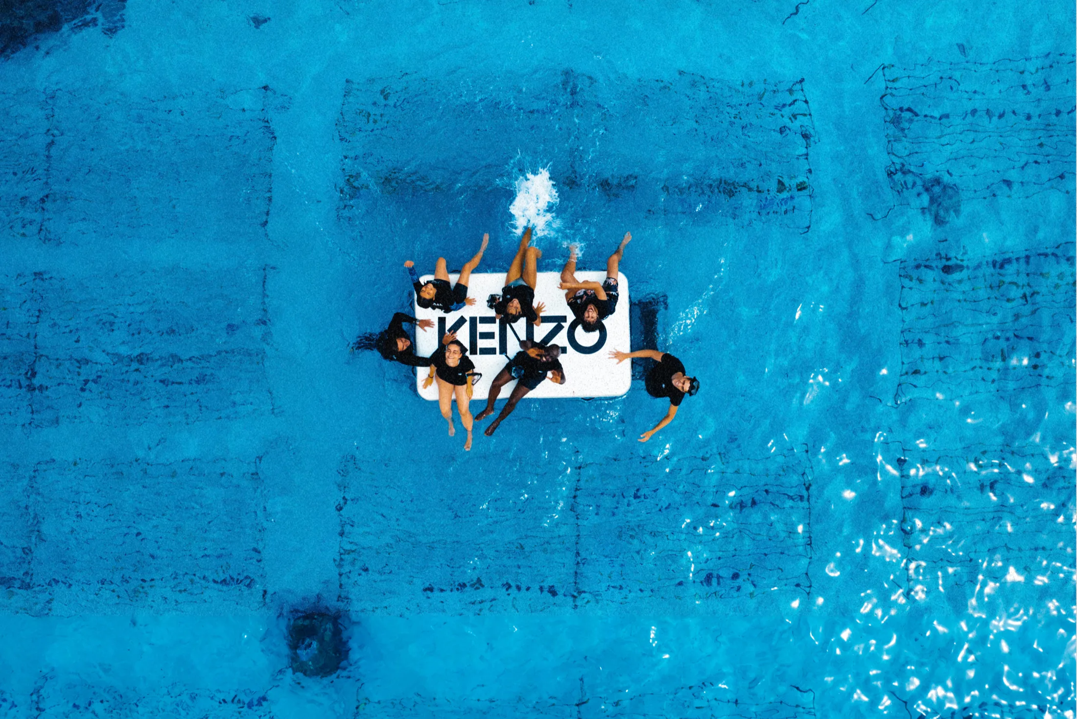 Swimmers around a floating Kenzo sign, seen from above