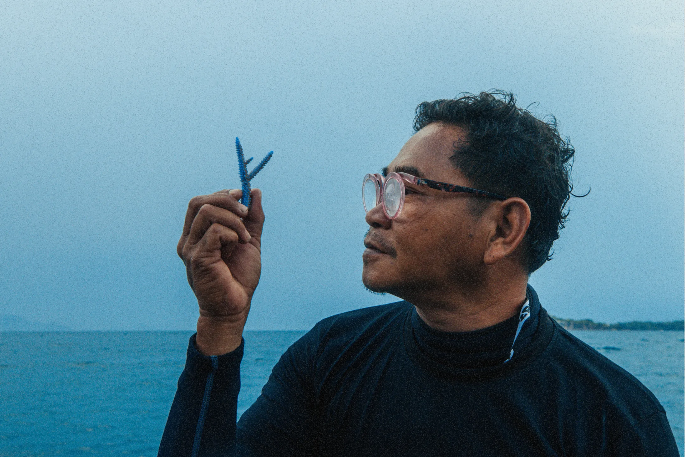 A Gardener at the water's edge holding a blue coral fragment up to the light