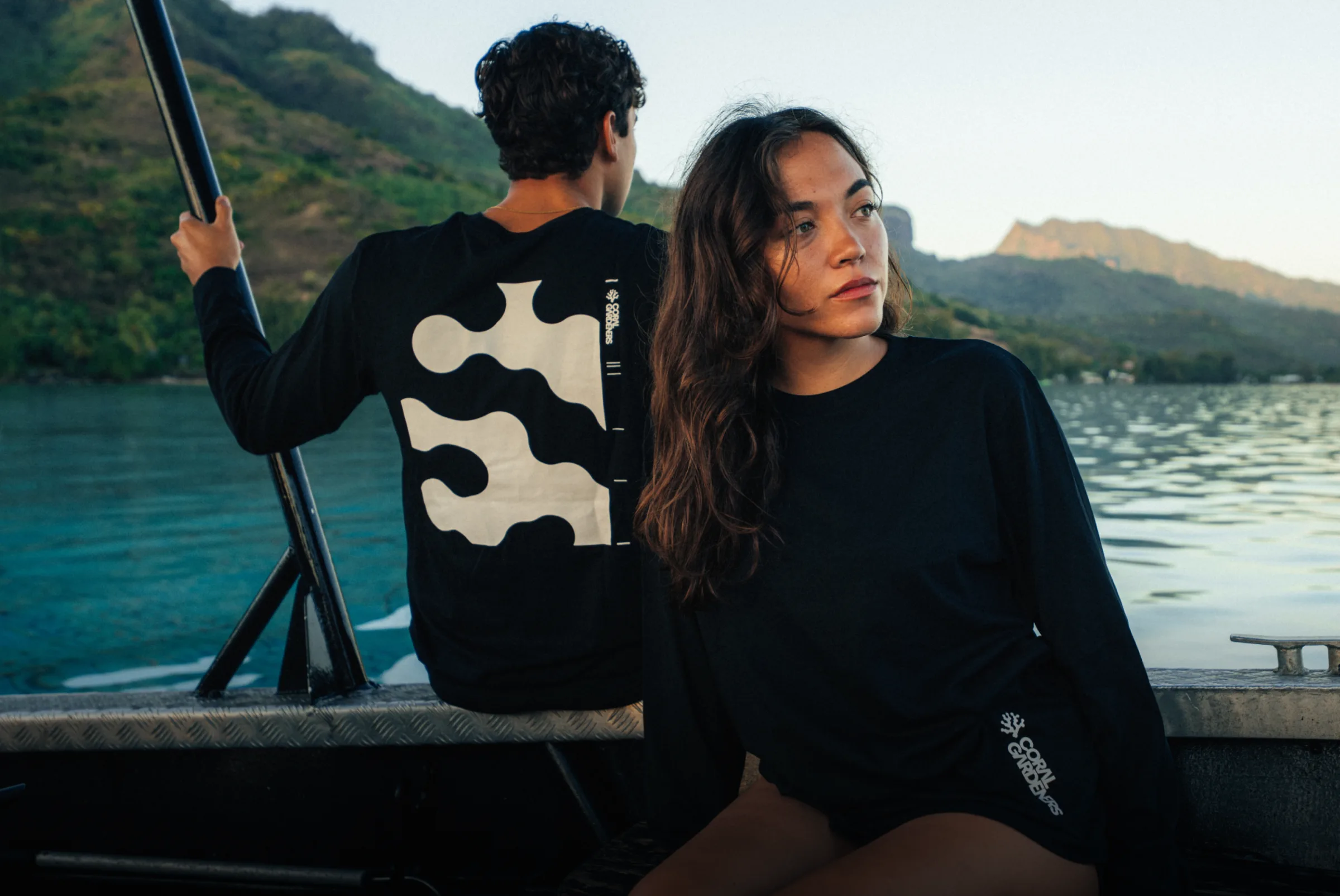 Two Gardeners in black Coral Gardeners long-sleeves on a boat in the lagoon, mountains behind